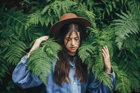 stylish hipster girl in hat sitting in fern bushes, among fern leaves in forest. creative portrait...