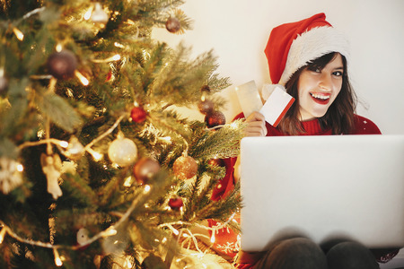 beautiful young woman in santa hat holding credit cards and sitting with laptop at golden beautiful...