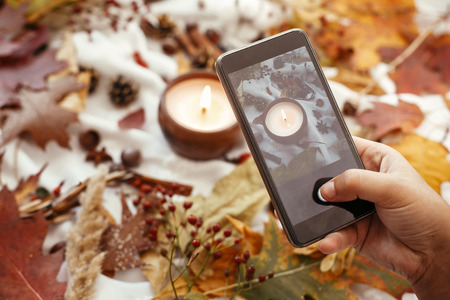 Hand holding phone and taking photo of stylish autumn flat lay of fall leaves, candle, berries,...