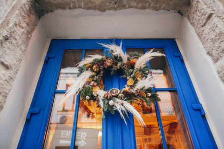 Modern christmas wreath with oranges,herbs, rustic ornaments, pine cones, branches on blue door in european city street. Festive decorations and illumination in city center,の写真素材