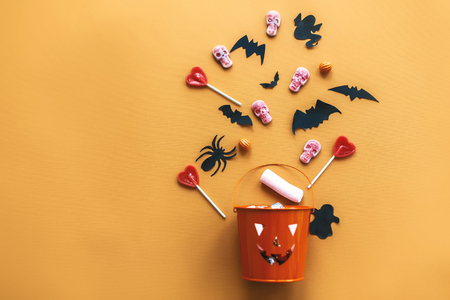 Halloween Jack o Lantern bucket with holiday candy, bats,spiders, skulls on orange paper, flat lay. Space for text. Halloween background. Season's greeting card mockupの写真素材