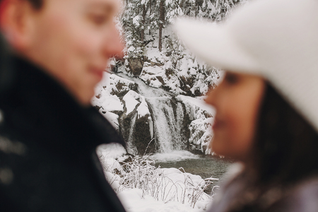 Stylish couple in love embracing in winter snowy mountains. Portraits of happy romantic man and woman smiling. Focus on waterfall in snow. Holiday getaway together.の写真素材