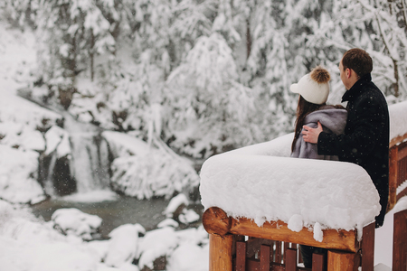 Stylish couple hugging and looking at snowy woods from wooden porch in mountains. Happy family relaxing on background of winter mountains. Holiday getaway together. Space for textの写真素材