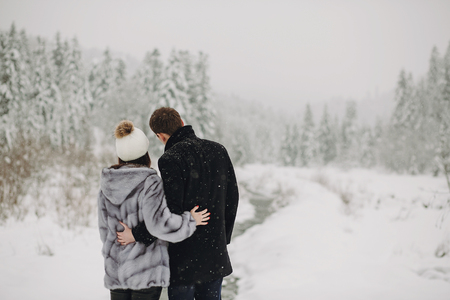 Stylish couple in love hugging  in snowy mountains. Happy family gently embracing in winter mountains and forest. View from back. Space for text. Holiday getaway togetherの写真素材