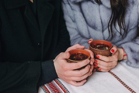 Stylish couple holding cups with hot tea in hands on wooden porch in winter snowy mountains. Happy romantic family with drinks. Holiday getaway together. Romantic momentsの写真素材