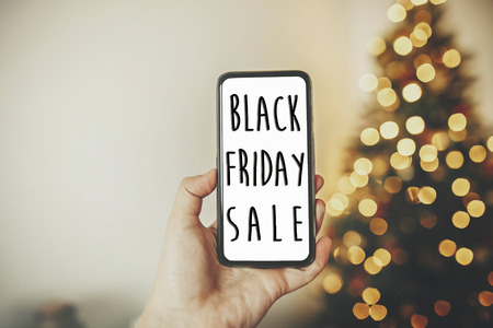 Black Friday big sale text. Holiday discount offer. Hand holding phone on background of golden beautiful christmas tree with lights in festive room.の写真素材