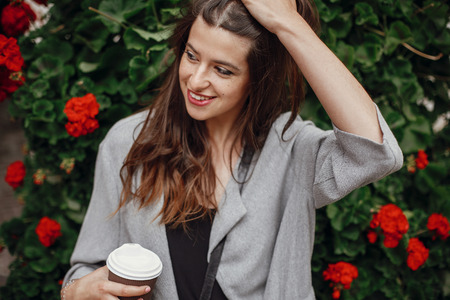 Gorgeous young woman holding cup of coffee and smiling in city street on background of flowers. Stylish happy hipster girl with beautiful hair enjoying time at cafe terraceの写真素材