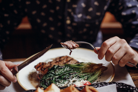 Man eating delicious juicy steak with grilled cabbage,tomatoes and cheese on table at cafe terrace in city street. Hands holding fork and knife and tasting bbq with vegetables in restaurantの写真素材
