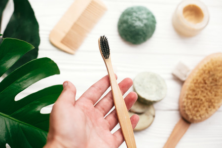 Hand holding natural bamboo toothbrush on background of shampoo bar, brush, toothpaste on white wood...