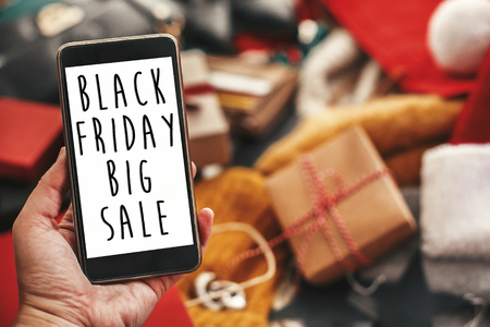 Black Friday big sale text on phone screen. Special discount christmas offer sign. Big Sales. Hand holding phone with advertising message at credit cards, bags, clothes, gifts.の写真素材