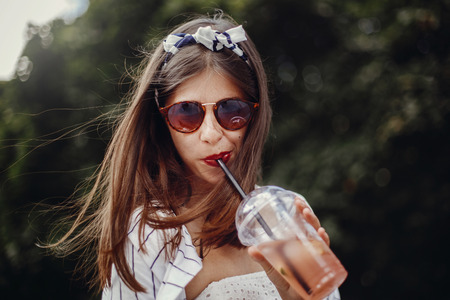 Happy hipster girl with sunglasses, in retro dress and headband, holding plastic cup and straw,...