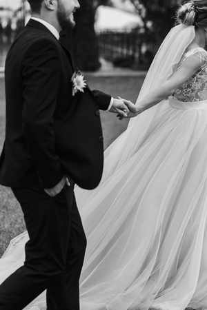 Gorgeous bride and stylish groom holding hands and walking outdoors. Happy Sensual wedding couple embracing. Romantic moments of newlyweds. Wedding photoの写真素材