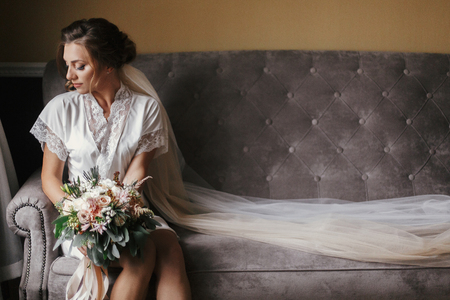 Gorgeous beautiful bride in silk robe and veil holding modern bouquet and sitting on stylish gray sofa. Happy stylish Bride with perfect makeup posing near window in soft light in the morningの写真素材