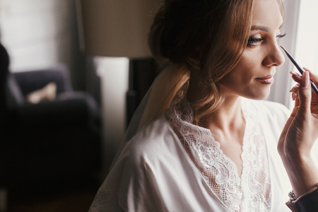 Stylish bride getting her make-up in the morning at window light. Makeup artist doing brows and applying eyeshadow on bride face close-up.の写真素材