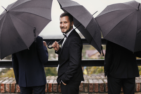 Stylish groom and groomsmen standing under black umbrella and posing. Confident man in suit holding umbrella in rainy outdoors. Rich Businessman. Finance wealth and businessの写真素材