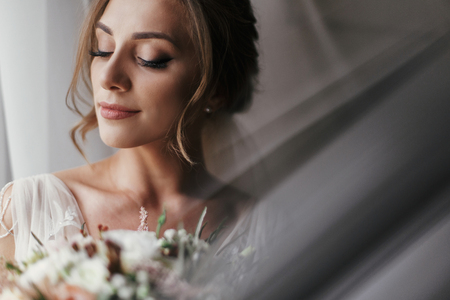 Gorgeous beautiful bride with bouquet posing at window in soft light. Happy Bride portrait in stylish gown, with perfect makeup, smiling and dreaming. Romantic moment. Space for textの写真素材