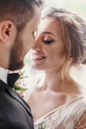 Gorgeous bride and stylish groom gently hugging and embracing at window light. Sensual wedding couple smiling in light. Happy romantic moments of newlyweds. Wedding photoの写真素材