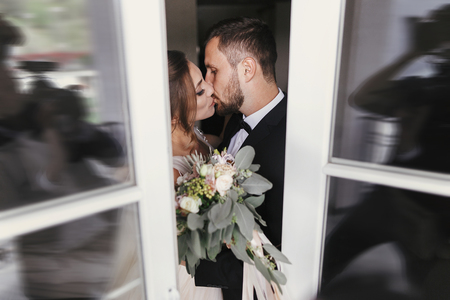 Gorgeous bride and stylish groom gently kissing at window. Sensual wedding couple embracing. Romantic moments of newlyweds. Creative wedding photo through glass. Copy spaceの写真素材
