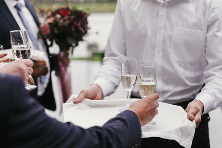 Waiter holding tray with glasses of champagne and serving them for toasts wedding reception. People cheering with alcohol drinks at party outdoors. Luxury Christmas feastの写真素材