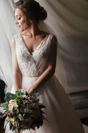 Gorgeous beautiful bride with bouquet posing at window in soft light. Happy  Bride in stylish gown and with perfect makeup smiling and dreaming. Bridal morning. Romantic momentの写真素材