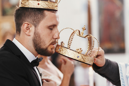 Bride and groom kissing golden crowns from priest hand during wedding ceremony. Spiritual couple. Wedding matrimony in church. Emotional romantic momentsの写真素材