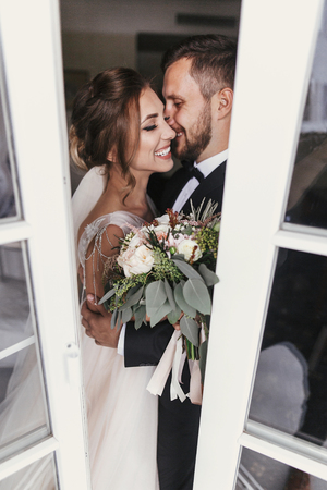 Gorgeous bride and stylish groom gently kissing at window. Happy wedding couple embracing. Romantic moments of newlyweds. Creative wedding photo through glass. Copy spaceの写真素材