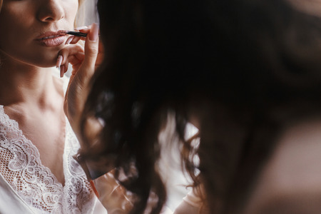 Stylish bride getting her make-up in the morning at window light. Makeup artist painting  lips with brush in nude lipstick on bride face close-up.の写真素材