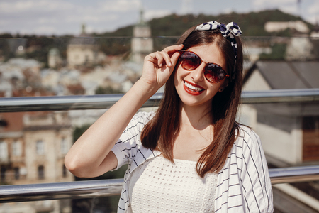 Beautiful stylish young woman in  sunglasses, retro dress and headband relaxing on roof in european city street. Hipster girl smiling and enjoying summer time in Lviv. Space for textの写真素材