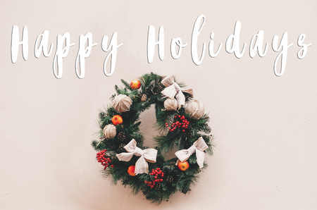 Happy Holidays text on stylish christmas wreath with red berries,bows and ornaments, pine cones, branches. Season's greetings card. Merry Christmas and Happy New Yearの写真素材