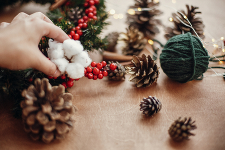Hand putting cotton on wreath,pine cones, fir branches, red berries, thread, cinnamon,  lights on rustic wooden background. Details for making christmas wreath at workshop. Atmospheric imageの写真素材
