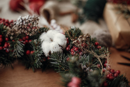 Details of rustic Christmas wreath, cotton close up. Fir branches with red berries,pine cones,ribbon, cinnamon on rustic wood. Atmospheric moody image at holiday workshopの写真素材