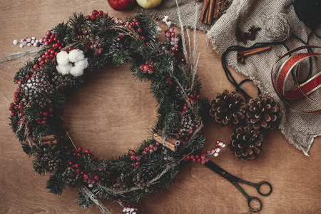 Rustic Christmas wreath, flat lay. Fir branches with red berries,pine cones,ribbon, scissors, cinnamon, cotton, apples on rustic wooden background. Atmospheric moody imageの写真素材