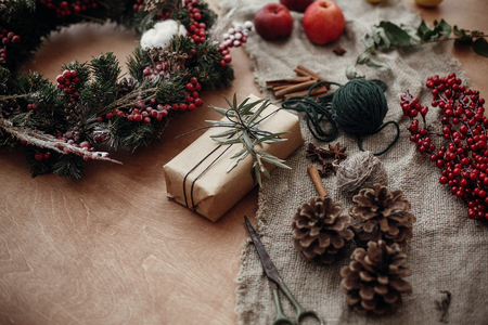Rustic Christmas wreath and stylish craft gift box with green branch at fir, red berries,pine cones,rope, cinnamon, scissors on rustic wood. Atmospheric moody image, happy winter holidayの写真素材