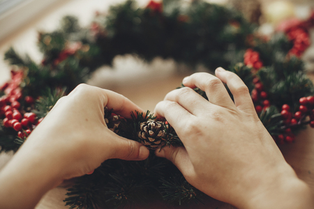 Hands making rustic christmas wreath, holding pine cone at fir branches, red berries ,  cotton on rustic wooden background. Atmospheric moody image at winter holiday workshopの写真素材
