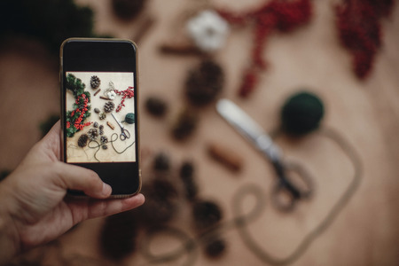 Hand holding phone and taking photo of rustic christmas wreath flat lay. Photo process of making christmas wreath at holiday workshop with fir branches,berries, cones. Copy spaceの写真素材