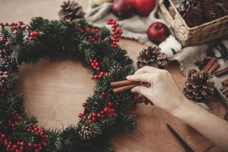 Hands making rustic christmas wreath, holding cinnamon at fir branches, red berries,pine cones,rope, scissors, cotton on rustic wooden background. Atmospheric moody image at holiday workshopの写真素材