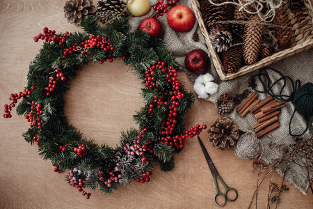 Rustic Christmas wreath, flat lay. Fir branches with red berries,pine cones,ribbon, scissors, cinnamon, cotton, apples on rustic wood. Atmospheric moody image at holiday workshopの写真素材