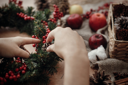 Rustic Christmas wreath. Hands holding fir branches, red berries and pine cones,rope, scissors, cinnamon, cotton on rustic wooden background. Atmospheric moody image at holiday workshopの写真素材