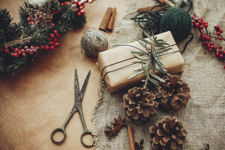 Details of rustic Christmas wreath. Fir branches with red berries,pine cones, cinnamon, scissors and craft gift box on rustic wood. Atmospheric moody image at winter holiday workshopの写真素材