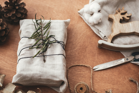 Zero waste  holidays.  Stylish christmas rustic gift wrapped in linen fabric with green branch on wooden table with pine cones, reindeer, scissors, twine, cotton. Simple eco presents plastic free.の写真素材
