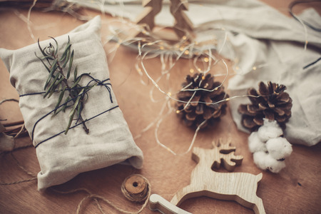 Merry Christmas, eco concept. Stylish rustic gift wrapped in linen fabric with green branch on wooden table with pine cones, reindeer, lights. Simple eco presents. Zero waste  holidays.の写真素材