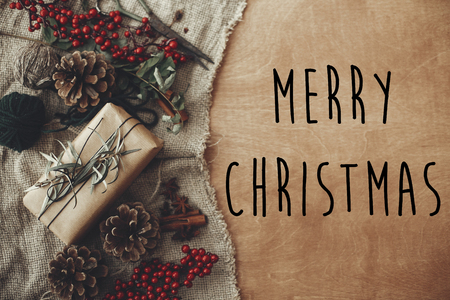 Merry Christmas text sign on stylish rustic christmas gift box with fir branches, red berries, pine cones on rustic wood. Atmospheric image. Seasonal greeting cardの写真素材