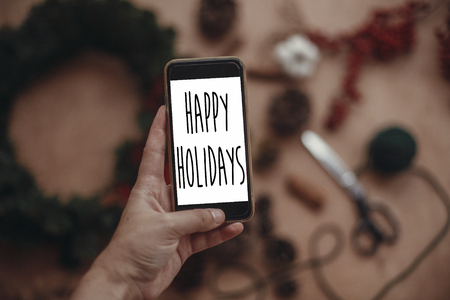 Happy Holidays text sign on phone screen in hand on background of rustic christmas wreath, fir branches,red berries, pine cones. Seasonal greeting card. Atmospheric image.の写真素材