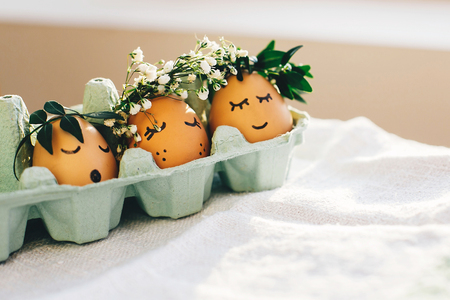 Happy Easter,  rustic eco concept. Stylish easter eggs in floral wreaths and  cute faces in carton tray. Modern cute easter eggs with flowers crown and sleepy eyes in sunny light. Space for textの写真素材