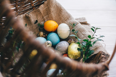 Stylish easter eggs on cloth in rustic basket on white wooden background. Easter hunt concept. Modern easter eggs painted with natural dye and green buxus branches. Happy Easterの写真素材