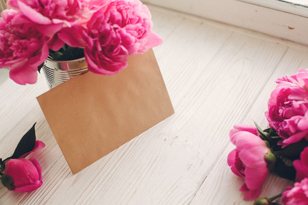 lovely pink peonies bouquet with empty craft card on rustic white wood at old window in light, top view, space for text. floral greeting card mock-up. happy mothers day conceptの写真素材