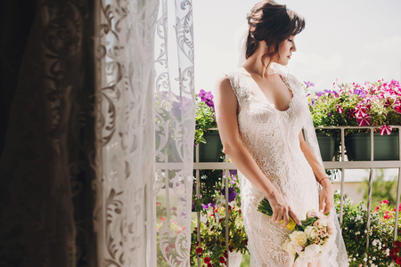Gorgeous bride in amazing gown and with wedding bouquet posing on balcony in flowers and sunny light. Beautiful woman getting ready in the morning. Wedding preparationsの写真素材