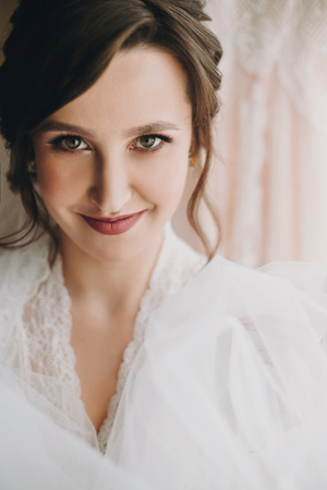 Beautiful stylish brunette bride posing in silk robe under veil in the morning. Happy woman model with perfect makeup and hairstyle, getting ready for wedding dayの写真素材