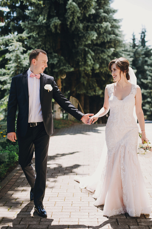 Gorgeous bride in amazing gown and stylish groom walking and smiling in sunny city street. Beautiful happy wedding couple enjoying time in sunny outdoors. Romantic momentsの写真素材