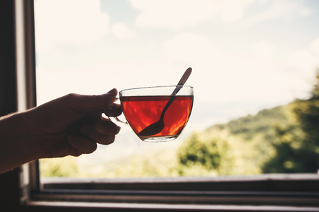 Hand holding hot tea in glass cup on background of  on beautiful landscape of woods and mountains. View from wooden window. Space for text. Travel and vacationsの写真素材